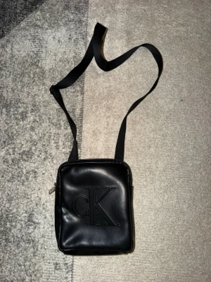 Svart axelväska från Calvin Klein - Säljer en stilren svart axelväska från Calvin Klein med präglad CK-logga på framsidan. Väskan har en fyrkantig form, justerbar axelrem och är tillverkad i slätt syntetmaterial. Perfekt för att bära dina essentials.