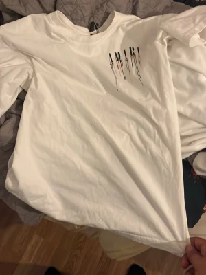 Vit AMIRI t-shirt med tryck - Säljer en vit t-shirt från AMIRI med coolt tryck av märkesnamnet och färgstänk på både bröstet och ryggen. T-shirten har rund hals och korta ärmar, perfekt för en trendig och avslappnad stil.