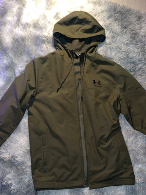 Olivgrön vindjacka från Under Armour - Snygg olivgrön vindjacka från Under Armour med huva och dragsko. Jackan har hel dragkedja framtill, två sidofickor och meshfoder på insidan för extra komfort. Perfekt för blåsiga dagar och sportiga outfits.