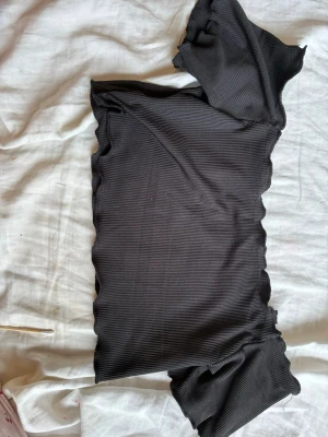 Svart ribbad oneshoulder topp - Säljer en svart ribbad oneshoulder topp med vågiga kanter. Toppen är tight och har en modern, asymmetrisk design som ger en cool vibe. Perfekt att styla med jeans eller kjol för en trendig look.