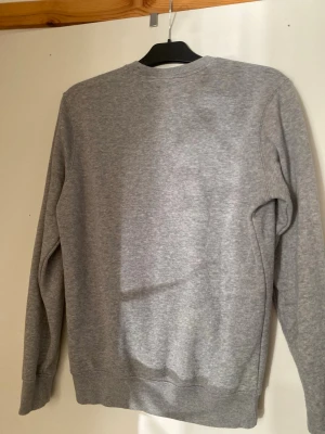 Grå sweatshirt från Champion - Klassisk grå sweatshirt från Champion med rund halsringning och diskret logga broderad på bröstet. Tröjan har långa ärmar och ribbade muddar vid ärmslut och nederkant. Perfekt för en avslappnad och sportig stil.