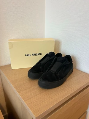 Svarta sneakers från Axel Arigato - Stilrena svarta sneakers från Axel Arigato i mocka med minimalistisk design och tjock sula. Skorna har diskret logga på sidan och en klassisk låg siluett som passar till det mesta. Perfekta för dig som gillar en clean och modern look.