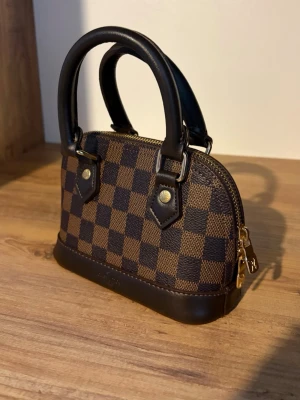 Brun handväska med axelremsband - Snygg handväska i klassiskt brunt damier-mönster. Väskan har två bruna handtag, guldfärgade metalldetaljer och dragkedja. Tillverkad i canvas med läderdetaljer. Perfekt accessoar för dig som gillar lyxig och tidlös stil. Axelremsband följer med.