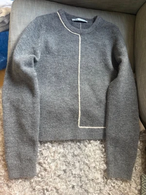 Grå stickad tröja med vit detalj - Mysig grå stickad tröja med lång ärm och en unik vit linje som går från halsen ner över bröstet och runt till ryggen. Tröjan har rund halsringning och ribbade muddar vid ärmslut och nederkant.