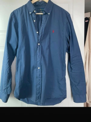 Blå skjorta från Ralph Lauren - Säljer en klassisk blå skjorta från Ralph Lauren i slim fit. Skjortan har långa ärmar, knappar framtill och den ikoniska röda loggan broderad på bröstet. Tillverkad i lätt bomullstwill som känns skön mot huden. Perfekt för en clean och stilren look.