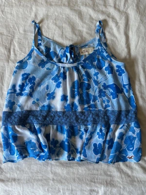 Blommig blå linnetopp från Hollister - Söt linnetopp från Hollister med blått blommönster på vit botten. Toppen har tunna axelband, spetsdetalj i midjan och knytning i ryggen. Perfekt för varma dagar och ger en fräsch sommarkänsla.