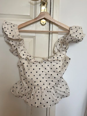 Vit prickig blus med volanger - Supersöt vit blus med svarta prickar och volangdetaljer längs axlarna. Toppen har en öppen rygg med knytband och en lätt peplum nedtill. Perfekt för dig som vill ha en lekfull och trendig look.