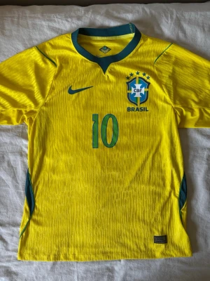 Brasilien Neymar JR matchtröja gul - Säljer en gul Brasilien fotbollströja från Nike med Neymar JR och nummer 10 på ryggen. Tröjan har gröna detaljer, CBF-emblem och Nike-logga på bröstet. Tillverkad i lätt och ventilerande material, perfekt för match eller träning.