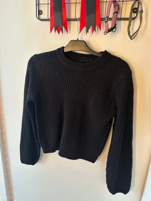 Svart stickad tröja - En svart stickad tröja med rund halsringning och långa ärmar. Tröjan har ett grovt stickat mönster och ribbade muddar vid ärmslut och nederkant. Perfekt för kalla dagar när du vill vara både varm och stilren.