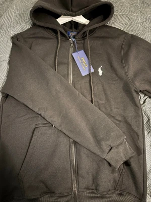 Svart hoodie från Polo Ralph Lauren - Svart hoodie med dragkedja från Polo Ralph Lauren. Klassisk design med huva och dragsnören, två fickor framtill och den ikoniska vita logotypen broderad på bröstet. Tillverkad i mjukt bomullsmaterial som är skönt att bära.