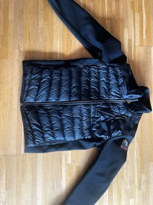 Svart pufferjacka från Parajumpers - Snygg svart pufferjacka från Parajumpers med quiltad front och släta ärmar. Jackan har dragkedja framtill, hög krage och en patch på ena ärmen. Materialet är en mix av glansig nylon och strukturerad textil på ärmarna. Perfekt för kalla vinterdagar.