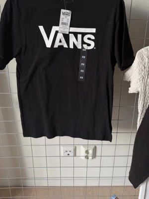 Svart Vans t-shirt med logga - Svart t-shirt från Vans med stor vit logga framtill. Klassisk passform och rund halsringning. Tillverkad i mjuk bomull som känns skön mot huden. Perfekt för dig som gillar streetwear och enkel stil.