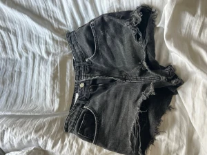 Svarta jeansshorts - Säljer ett par svarta jeansshorts med råa, fransiga kanter och slitningar. Shortsen har klassisk femficksmodell och knappgylf. Perfekta för en avslappnad och trendig look.