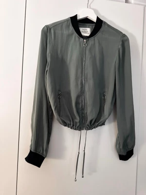Militär grön  bomberjacka från Bershka - Snygg bomberjacka från Bershka med svarta muddar och krage. Jackan har dragkedja framtill, två fickor med dragkedja och dragsko i midjan för justerbar passform. Perfekt för en trendig och avslappnad stil.