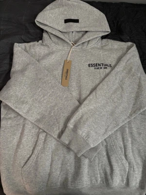 Grå Essentials Fear of God hoodie - Snygg grå hoodie från Essentials Fear of God med svart logga på bröstet. Klassisk huva, känguruficka och ribbade muddar. Tillverkad i mjukt bomullsmaterial som är perfekt för en avslappnad streetwear-look.
