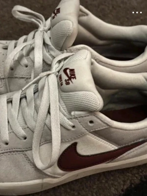 Nike SB vita sneakers med vinröd swoosh - Nike SB sneakers i vitt med vinröd swoosh på sidan och matchande logga på plösen. Skorna har lågt skaft, vita skosnören och en slitstark yttersula med mönster för bra grepp. Ovandelen är i mocka och mesh för skön känsla och stilren look.
