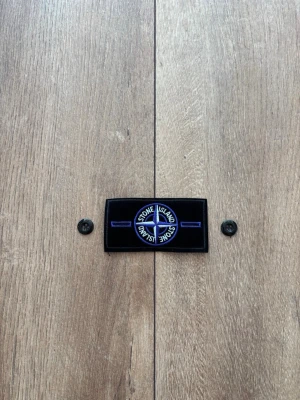 Stone Island Patch / Badge – Svart med blå/lila brodyr & knappar - Säljer en klassisk Stone Island-badge i svart med blå/lila brodyr, inklusive tillhörande originalknappar precis som på bilderna. Patchen är helt ny, oanvänd och i perfekt skick. Passar utmärkt som reservdel eller tillbehör till din Stone Island-jacka eller tröja. 