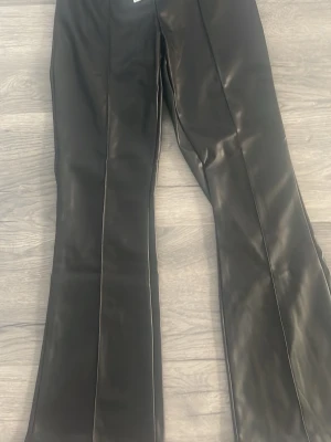 Svarta utsvängda fuskskinnjeans - Snygga svarta jeans i fuskskinn med utsvängda ben och markerad pressveck. Perfekta för dig som vill ha en edgy och trendig look. Materialet ger en cool glansig finish och byxorna har hög midja.