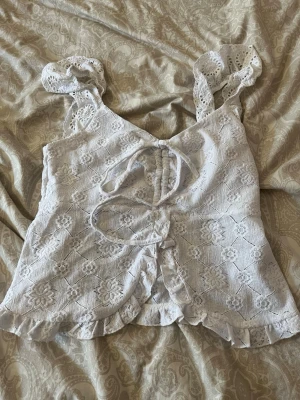 Vit spetslinnetopp med volang - Supersöt vit linnetopp med broderade blommor, volangkant och breda axelband i spets. Toppen har snörning framtill och en lätt croppad passform. Perfekt för sommarens alla tillfällen och ger en romantisk vibe.