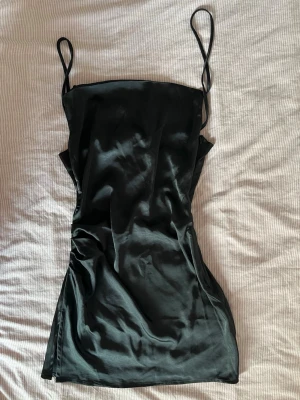 Svart satin kortklänning med smala band - Snygg svart kortklänning i satin med tunna axelband och rak urringning. Klänningen har en figurnära passform och ett stilrent, minimalistiskt uttryck. Perfekt för dig som gillar en enkel men elegant look.