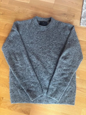 Grå stickad tröja från Jack & Jones - Mysig grå stickad tröja från Jack & Jones Premium med rund halsringning och ribbade muddar vid ärmslut och nederkant. Tröjan har en fluffig och mjuk struktur som gör den perfekt för kyliga dagar. Enkel och stilren design som passar till det mesta.