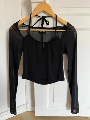 Svart mesh topp med knytband - Säljer en svart croppad topp med meshärmar och knytband i nacken. Toppen har en liten v-ringning framtill och är tillverkad i ett tunt, syntetiskt material. Perfekt för dig som gillar trendiga detaljer och en edgy look.
