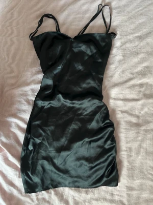 Svart satin kortklänning med snörning - Snygg svart kortklänning i satin med tunna axelband och trendig snörning i ryggen. Klänningen har en figurnära passform och en slät, glansig yta som ger en lyxig känsla. Perfekt för dig som vill sticka ut med en elegant och modern look. Använd endast en gång!