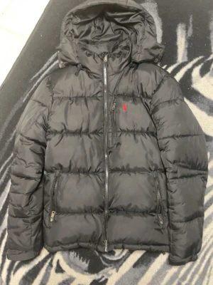 Svart pufferjacka från Polo Ralph Lauren - Säljer en svart pufferjacka från Polo Ralph Lauren med huva och dragkedja framtill. Jackan har två sidofickor med dragkedja och en bröstficka. Klassisk röd logga broderad på bröstet. Perfekt för kalla vinterdagar och riktigt skön att bära.