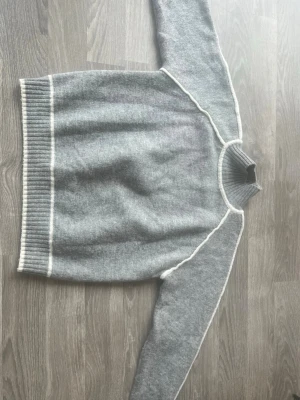 Grå stickad tröja med vita detaljer - Mysig grå stickad tröja med vita kontrastsömmar längs ärmar, axlar och halsringning. Tröjan har ribbstickade muddar vid hals, ärmslut och nederkant. Perfekt för kyliga dagar och enkel att matcha med jeans eller kjol.