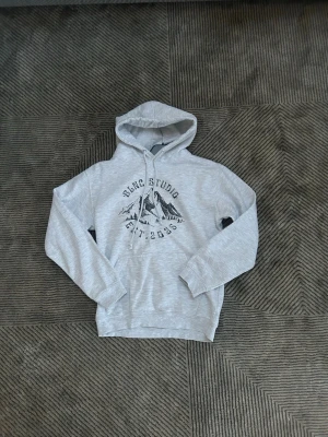 Ljusgrå hoodie från blnc studio  - Ljusgrå hoodie med huva från blnc studio  och dragsko. På bröstet finns ett tryck med texten 'BCNC Studio Est. 2003' och ett bergmotiv. Hoodien har ribbade muddar vid ärmar och nederkant. Perfekt för en avslappnad och chill stil.