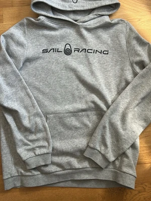Grå hoodie från Sail Racing - Snygg grå hoodie från Sail Racing med svart logga tryckt på bröstet. Klassisk känguruficka framtill och ribbade muddar vid ärmar och nederkant. Tillverkad i mjukt sweatshirtmaterial och har en stor huva för extra komfort.