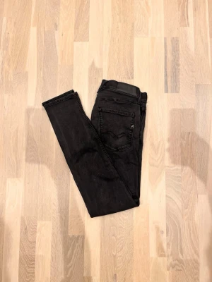 Svarta Replay 31/32 - Säljer ett par svarta skinny jeans från Replay med klassisk femficksdesign och diskret slitning. Jeansen har en snygg tvätt och Replay-logga på bakfickan samt läderpatch i midjan. Perfekta för en trendig och avslappnad stil.