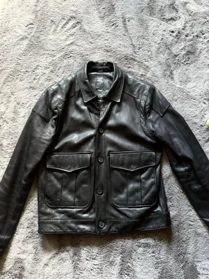 Svart skinnjacka från Emilio - Snygg svart skinnjacka från Emilio i storlek Medium. Jackan har klassisk krage, stora fickor med lock framtill och stängs med knappar. Tillverkad i äkta läder med en stilren och rak passform som ger en cool look.