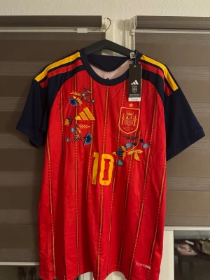 Spansk landslagströja Adidas röd - Röd och marinblå fotbollströja från Adidas med gula detaljer, broderade blommor och nummer 10 på framsidan. Tröjan har korta ärmar, rund hals och officiellt Spanien-emblem på bröstet. Tillverkad i lätt och ventilerande material.