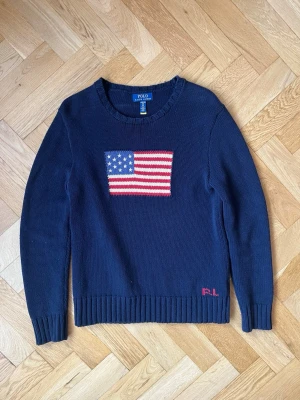 Mörkblå stickad tröja med USA-flagga - Mörkblå stickad tröja från Polo Ralph Lauren med amerikansk flagga på bröstet och diskret röd  RL brodyr vid nederkanten. Tröjan har rund halsringning, långa ärmar och ribbade muddar. Perfekt för dig som gillar klassisk preppy stil.