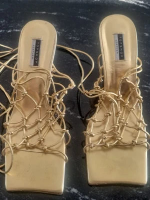 Guldiga klackskor från Topshop - Säljer ett par guldiga klackar från Topshop. Skorna har fyrkantig tå och långa snöre som ger en unik look. Perfekta för dig som vill sticka ut med ett par riktigt snygga och moderna klackar. Använt en gång bara. 