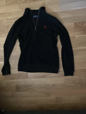 Svart halvzip tröja från Polo Ralph Lauren - Svart långärmad tröja med halv dragkedja och hög krage från Polo Ralph Lauren. Klassisk röd logga broderad på bröstet. Tröjan har ribbade muddar och är tillverkad i mjukt material som passar perfekt till en avslappnad stil.