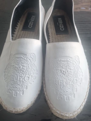 Vita espadrilles från Kenzo  - Säljer ett par vita loafers från Kenzo i läder med det ikoniska tigermotivet. Skorna har en klassisk espadrillesula i flätad jute och en stilren, rundad tå. Perfekta för dig som vill ha något trendigt och bekvämt. Har aldrig använt för att dem var för små. Storlek 38