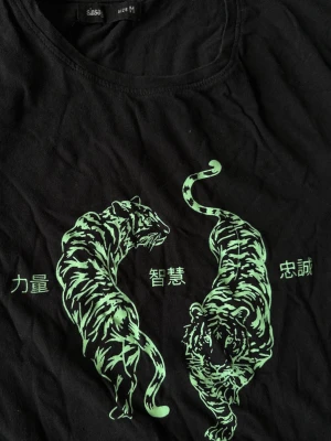 Svart t-shirt med gröna tigrar - Cool svart t-shirt med tryck av två gröna tigrar och asiatiska tecken på bröstet. T-shirten har rund hals och korta ärmar. Materialet känns mjukt och skönt, perfekt till vardags. Passar dig som gillar djurmotiv och streetstyle.