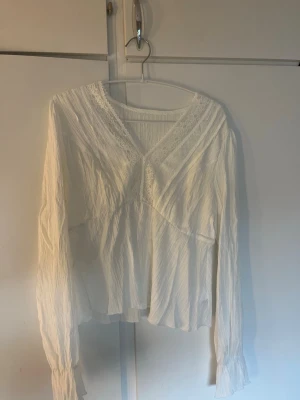 Vit blus med spetsdetaljer - Säljer en luftig vit blus med v-ringning och fina spetsdetaljer längs halsringningen. Blusen har långärmad design och är tillverkad i ett lätt, krinklat tyg som ger en avslappnad känsla. Perfekt att styla med jeans eller kjol.