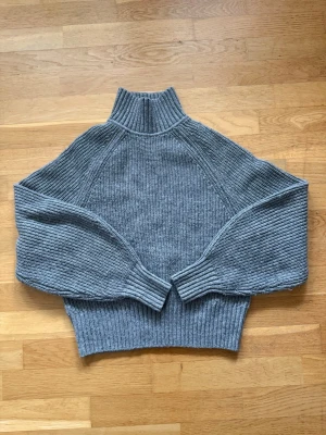 Grå stickad polotröja - Mysig grå stickad polotröja med bred ribbad mudd i hals, ärmslut och nederkant. Tröjan har en avslappnad passform och är tillverkad i ett mjukt, tjockt material som håller dig varm. Perfekt för kyliga dagar och enkel att styla med jeans eller kjol.