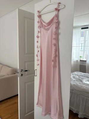 Ljusrosa långklänning med blommor - Säljer en elegant ljusrosa långklänning i satinliknande material. Klänningen har smala axelband med dekorativa blommor och en mjukt draperad urringning. Perfekt för dig som vill ha en romantisk och trendig look.