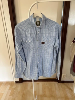 Vintage G-star Raw skjorta ⭐  - Snygg ljusblå skjorta med rutigt mönster och vita knappar. Skjortan har två bröstfickor med lock och knappstängning. Tillverkad i bomull och har lång ärm. Perfekt för en avslappnad men stilren look.
