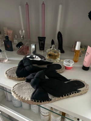 Svarta sandaler med flätad sula - Säljer ett par snygga sandaler med svart tyg och breda knutna band över foten. Sulan är flätad i naturfärgat material som ger en chill espadrille-vibe. Perfekta till sommaren och lätta att matcha med olika outfits.