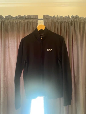 Svart zip hoodie från EA7 Emporio Armani - Snygg svart hoodie med dragkedja från EA7 Emporio Armani. Minimalistisk design med vitt EA7-tryck på bröstet och diskreta ränder i nacken på baksidan. Långärmad och tillverkad i mjukt material, perfekt för en clean och sportig look.