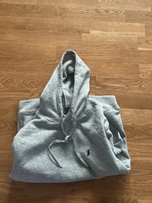 Grå hoodie från Polo Ralph Lauren - Snygg grå hoodie från Polo Ralph Lauren med klassisk huva och dragsko. Tröjan har den ikoniska lilla broderade loggan på bröstet och är tillverkad i mjukt material som känns skönt mot huden. Perfekt för en avslappnad och stilren look. Använd endast några få gånger o saknar defekter