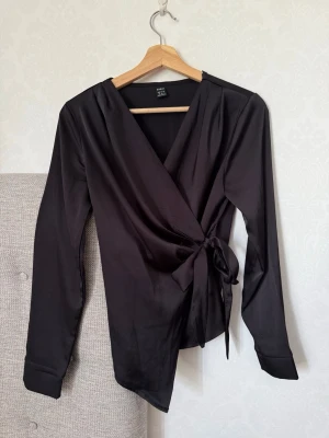 Svart omlottblus från SHEIN - Snygg svart omlottblus från SHEIN i glansigt, mjukt material. Blusen har långa ärmar, knytband i midjan och en djup V-ringning. Perfekt för dig som gillar stilrena och trendiga plagg med en elegant touch.