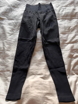 Svarta ribbade leggings med hög midja - Säljer ett par svarta leggings med ribbad struktur och hög midja. De är stretchiga och har en bred linning som ger extra stöd. Perfekta för en avslappnad och trendig look. Materialet känns mjukt och följsamt mot kroppen.