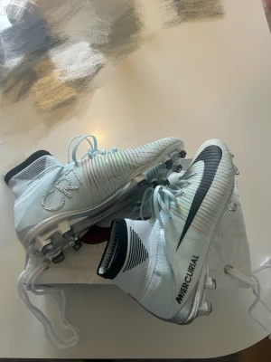 Nike Mercurial CR7 fotbollskor - Snygga fotbollsskor från Nike Mercurial-serien med CR7-detaljer. Skorna är vita med svarta och silverfärgade detaljer, högt skaft och snörning. Sulan har metallfärgade dobbar för bra grepp på planen. Perfekta för dig som vill sticka ut på matchen.