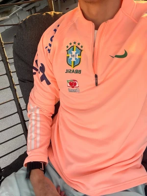 Rosa Brasil träningströja Nike - Säljer en snygg rosa långärmad träningströja från Nike med Brasil-logga och sponsormärken på bröstet och ärmarna. Tröjan har halv dragkedja framtill och grönt Nike-märke. Perfekt för fotbollsträning eller chill.